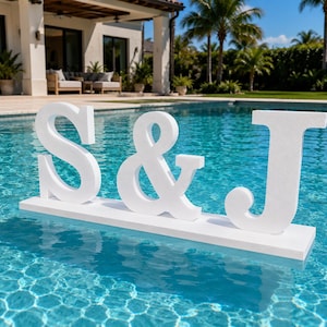 May include: A white foam sign featuring the letters S and J separated by an ampersand, mounted on a rectangular white base. The letters are uppercase with a serif font style and are positioned to float on the surface of a blue swimming pool.