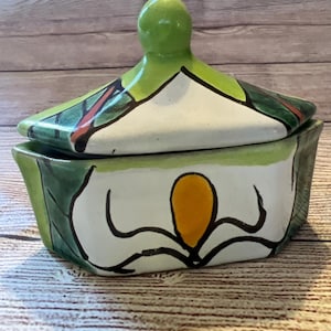 May include: A green, white and yellow ceramic lidded box with a floral design. The box has a geometric shape and a rounded lid.