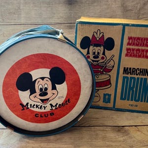 May include: Vintage Mickey Mouse Club marching drum with a red and white target design. The drum has a blue rim and a blue strap. The box next to the drum says "Disney on Parade Marching Drum."