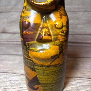 May include: A ceramic vase with a face-like design. The vase features a color palette of yellow, brown, and orange. The design includes a nose, eyes, and a mouth. The vase has a cylindrical shape and is likely used for decorative purposes.