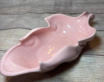 Può includere: Un piatto a forma di foglia in ceramica rosa chiaro. Il piatto ha una finitura liscia e lucida e una ciotola poco profonda. I bordi del piatto sono delicatamente curvi e hanno un aspetto increspato. Il piatto è probabilmente vintage e adatto per servire snack o come elemento decorativo.