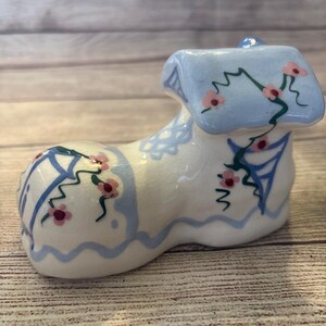 May include: A ceramic shoe-shaped figurine, painted white with blue and pink floral accents. The shoe has a light blue roof and decorative blue trim. The design includes pink flowers and green stems, adding a whimsical touch to the piece.