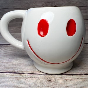 May include: White ceramic mug with a red smiley face. The mug has a large handle and a rounded shape.