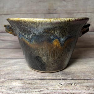 May include: A handmade ceramic bowl with two handles. The bowl has a dark gray glaze with streaks of brown, blue, and white.