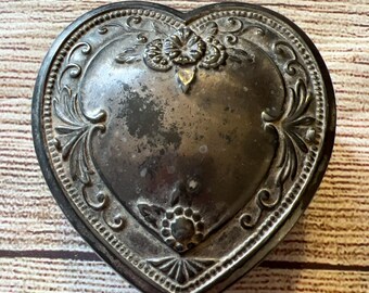 Puede incluir: Una caja de corazón de plata con un diseño ornamentado. La caja tiene un corazón en relieve en el centro y está decorada con motivos florales.