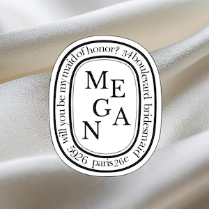 Puede incluir: Un diseño ovalado en blanco y negro con el nombre "Megan" en el centro. El diseño está rodeado de texto, incluyendo "Will you be my maid of honor?" y "34 boulevard bridesmaid 5926 paris 26e". El fondo es de tela blanca.