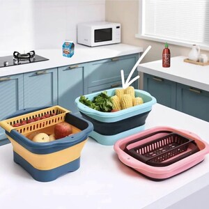 May include: Collapsible kitchen containers in blue, yellow, and pink. One container holds corn and lettuce, another has apples and tomatoes, and the third is empty. A microwave and stove are in the background.