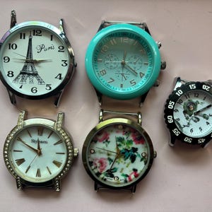May include: Five assorted wristwatches. One watch features the Eiffel Tower and the word "Paris". Another has a turquoise bezel. A third watch has a floral design. The fourth watch has a crystal-studded bezel. The fifth watch has a black bezel.