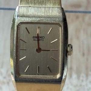 May include: A gold-tone Seiko Quartz wristwatch featuring a rectangular case and a matching integrated metal link bracelet. The dial is a metallic gold color with simple gold-tone hour markers, thin hands, and the text Seiko Quartz printed.
