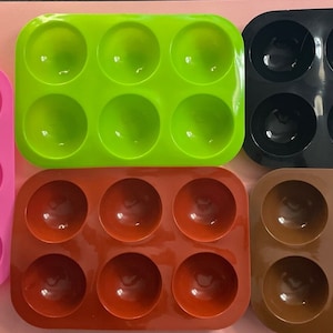 May include: Four silicone molds for making round treats. The molds are pink, green, black, and brown. Each mold has six round cavities.