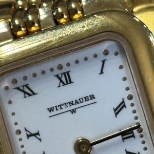 May include: Close-up of a gold-toned Wittnauer watch. The watch face is white with black Roman numerals and hands. The brand name is printed in black. The watch band is also gold-toned.