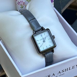 May include: A Laura Ashley wristwatch with a square face, white dial, and dark gray metallic case. The watch features a textured gray leather strap and is presented on a white cushion inside a branded box with the text Laura Ashley Since 1953.
