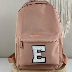 Könnte beinhalten: Ein rosafarbener Rucksack mit einem weißen Filzbuchstaben E auf der vorderen Tasche. Der Rucksack hat eine Reißverschlusstasche vorne und verstellbare Träger.