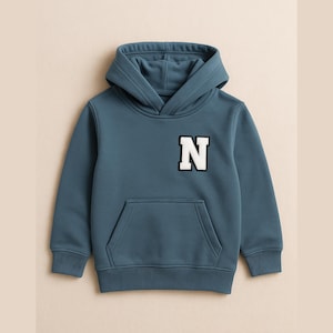 Könnte beinhalten: Ein tealblauer Kapuzenpullover mit Kängurutasche und einem weißen und schwarzen Buchstaben "N"-Aufnäher auf der Brust. Der Pullover ist aus einem weichen Material gefertigt und für Kinder konzipiert.