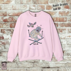 May include: A light pink sweatshirt with a whimsical illustration of a yeti skiing. The yeti is light blue and white, wearing skis and holding ski poles. Above the yeti is a skull wearing a wizard hat with wings. The text "hand drawn not AI" is printed on the top right.