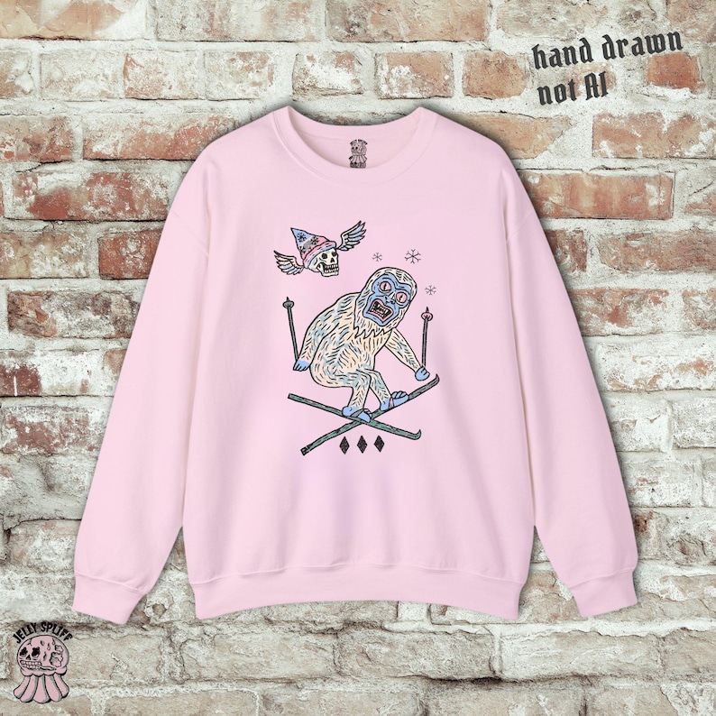 May include: A light pink sweatshirt with a whimsical illustration of a yeti skiing. The yeti is light blue and white, wearing skis and holding ski poles. Above the yeti is a skull wearing a wizard hat with wings. The text "hand drawn not AI" is printed on the top right.