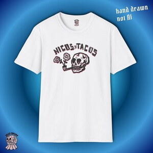 May include: White t-shirt featuring a graphic design with the text "NICOS TACOS" above a skull smoking a pipe, with flowers. The words "hand drawn not AI" are in the upper right corner. The shirt has a crew neck and short sleeves.
