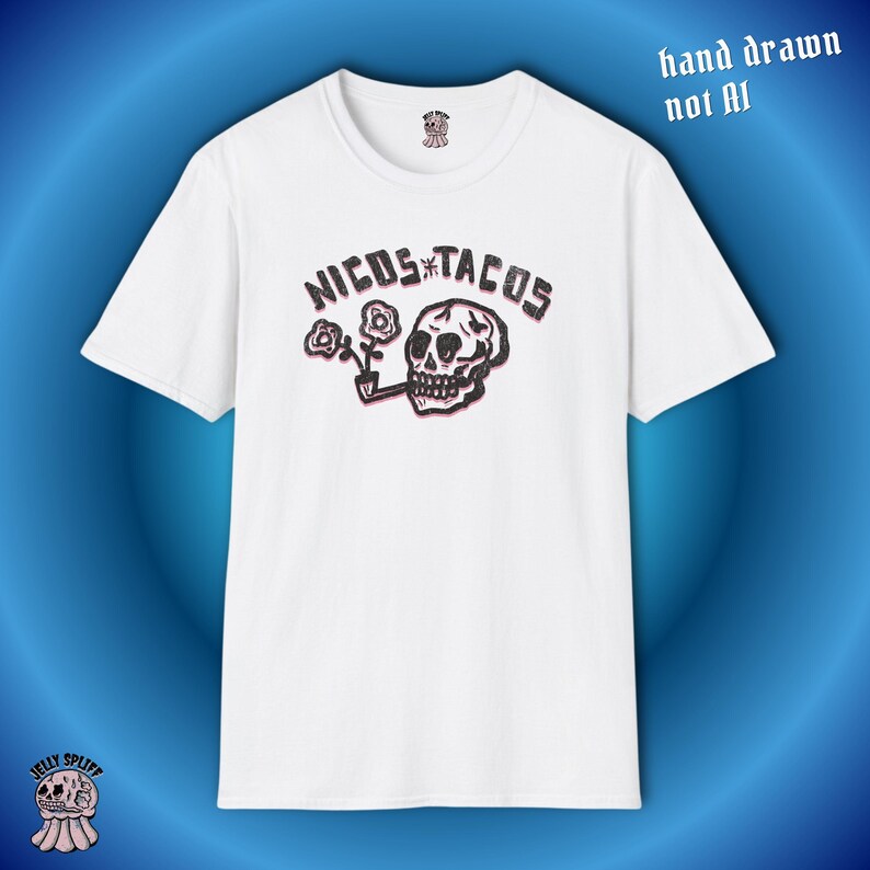 May include: White t-shirt featuring a graphic design with the text "NICOS TACOS" above a skull smoking a pipe, with flowers. The words "hand drawn not AI" are in the upper right corner. The shirt has a crew neck and short sleeves.