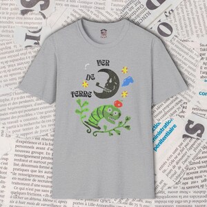 May include: Gray t-shirt with a whimsical graphic featuring a skull moon smoking a pipe, a green worm-like creature, and the text "VER DE TERRE". The design includes stars and a red apple.