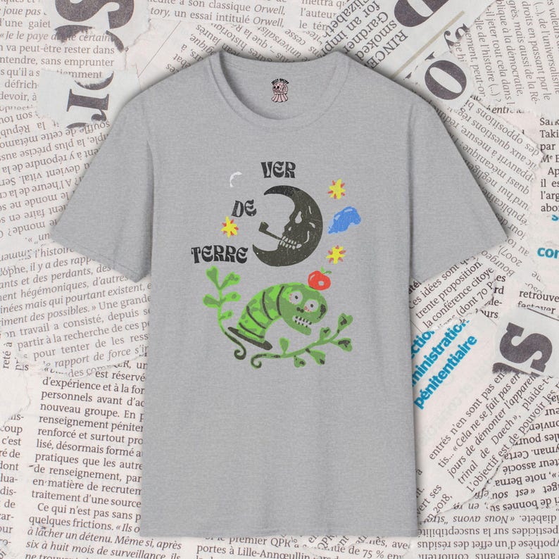May include: Gray t-shirt with a whimsical graphic featuring a skull moon smoking a pipe, a green worm-like creature, and the text "VER DE TERRE". The design includes stars and a red apple.