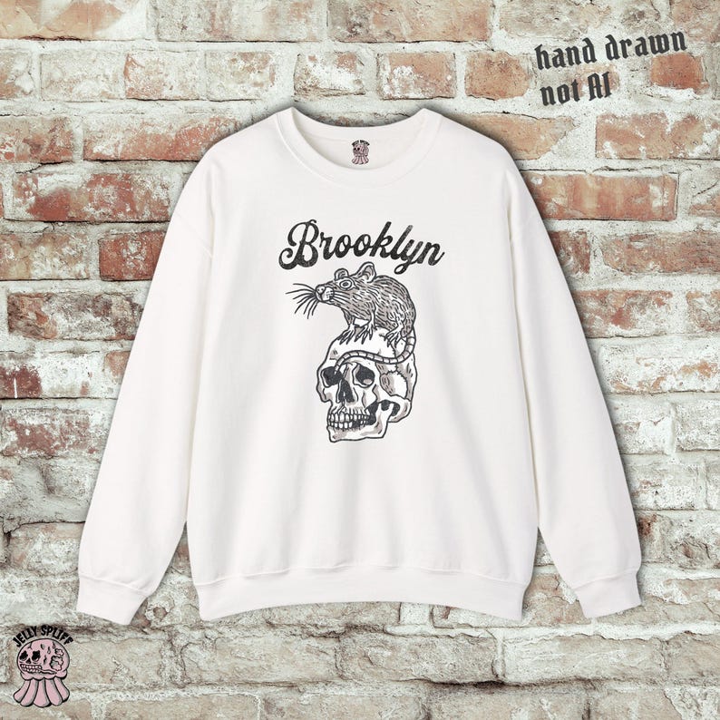 May include: White sweatshirt with a black graphic of a rat atop a skull, with the word "Brooklyn" above the graphic. The text "hand drawn not AI" is printed in black on the top right.