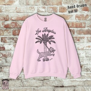 May include: Pale pink sweatshirt featuring a black illustration of a dog chained to a palm tree, with the words "Los Angeles" above. The text "hand drawn not AI" is in the upper right corner.