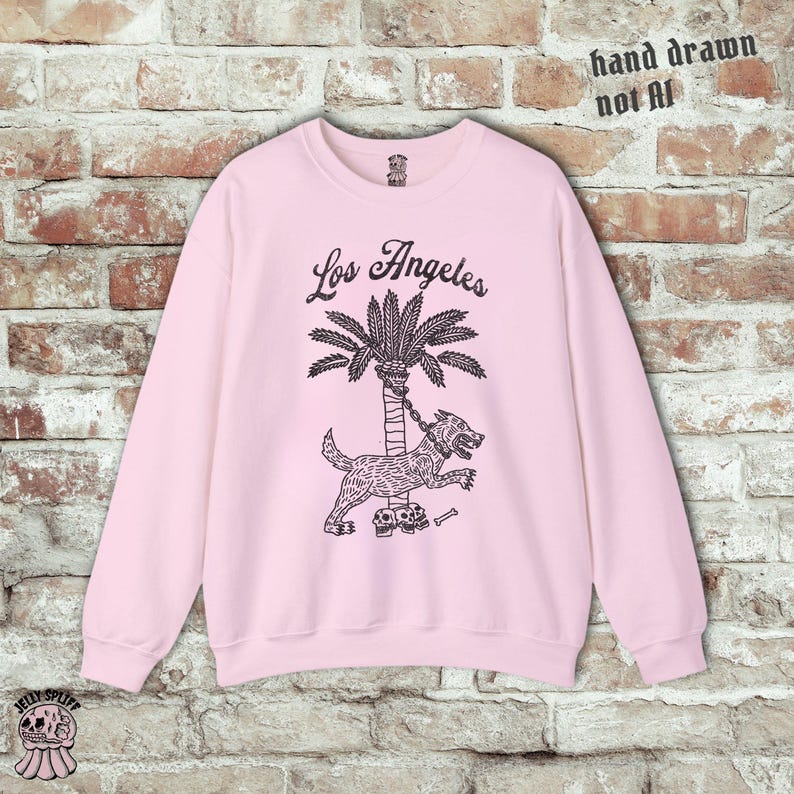 May include: Pale pink sweatshirt featuring a black illustration of a dog chained to a palm tree, with the words "Los Angeles" above. The text "hand drawn not AI" is in the upper right corner.