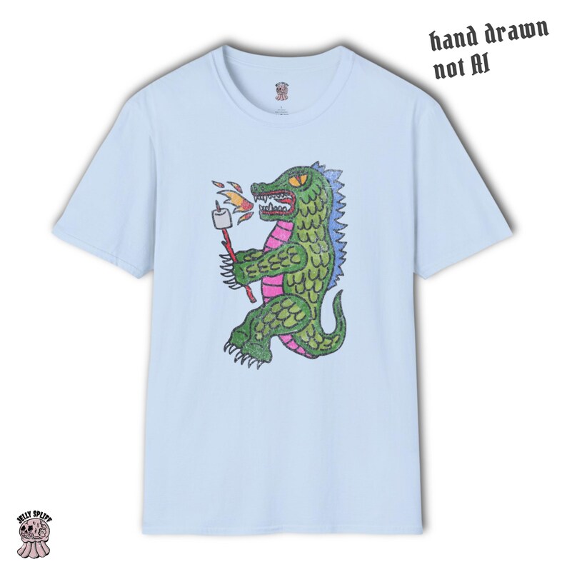 May include: Light blue t-shirt featuring a cartoon alligator roasting a marshmallow over a fire. The alligator is green, pink, and blue, with a marshmallow on a stick. Text on the shirt reads "hand drawn not AI".