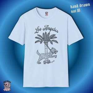 May include: A pale blue t-shirt with a black illustration of a dog chained to a palm tree, and the words "Los Angeles" above. The text "hand drawn not AI" is also visible. The shirt has a crew neck and short sleeves.