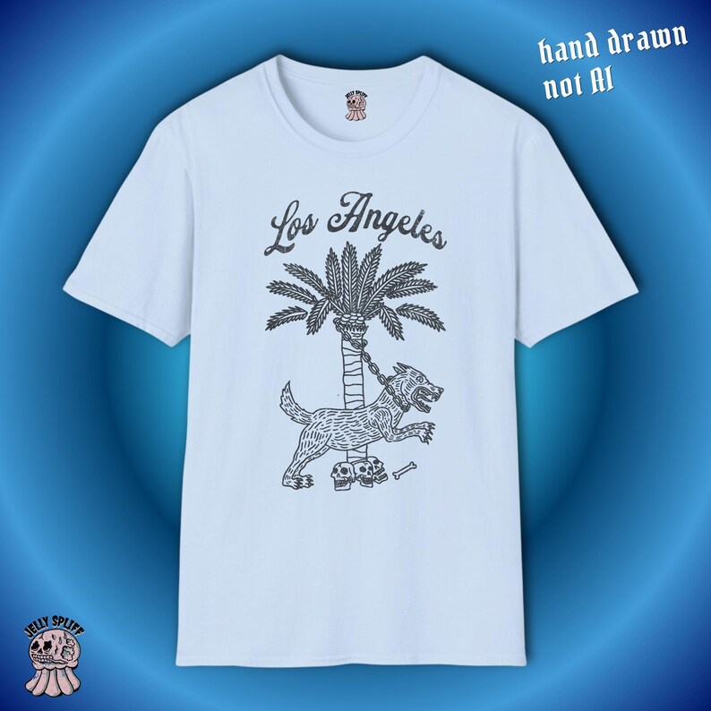 May include: A pale blue t-shirt with a black illustration of a dog chained to a palm tree, and the words "Los Angeles" above. The text "hand drawn not AI" is also visible. The shirt has a crew neck and short sleeves.