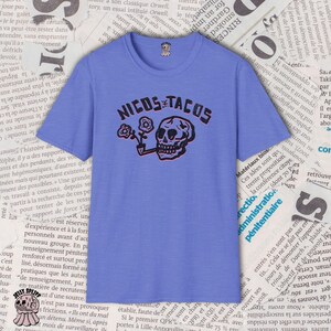 May include: A periwinkle blue t-shirt with the text "NICOS TACOS" in black and red, featuring a skull smoking a pipe and holding flowers. The shirt has a crew neck and short sleeves.