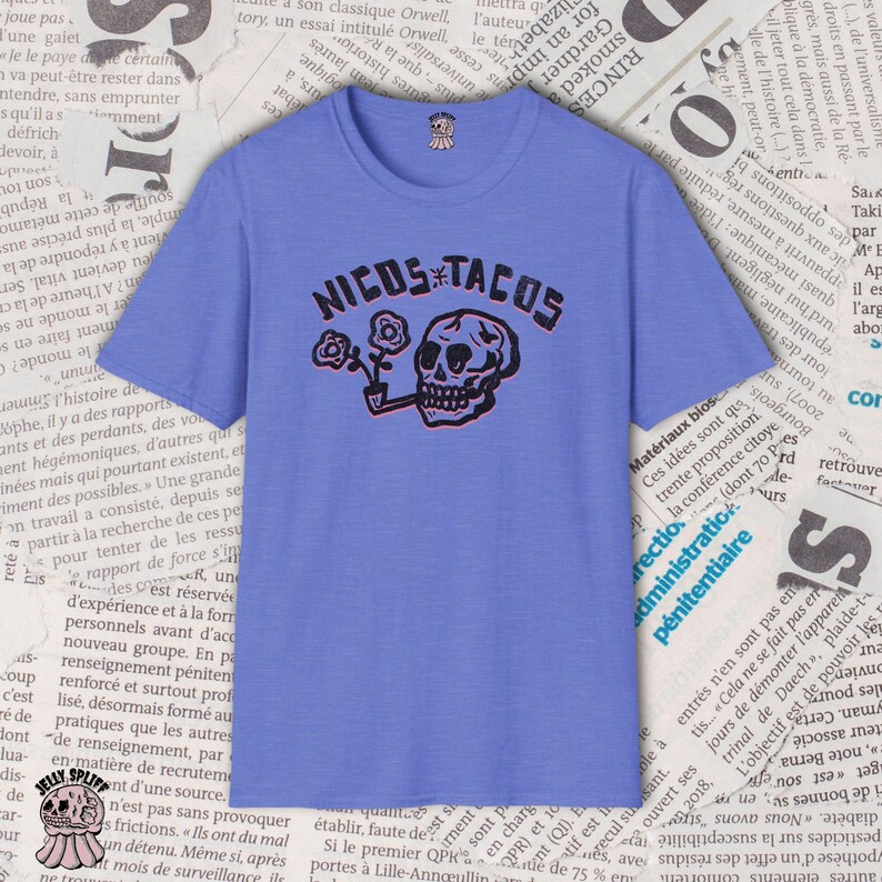 May include: A periwinkle blue t-shirt with the text "NICOS TACOS" in black and red, featuring a skull smoking a pipe and holding flowers. The shirt has a crew neck and short sleeves.
