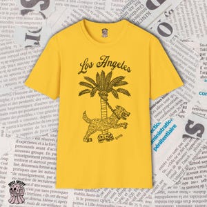 May include: Mustard yellow t-shirt with the words "Los Angeles" above a palm tree and a dog illustration. The dog is chained to the tree and has skulls at its feet. The shirt has a round neck.