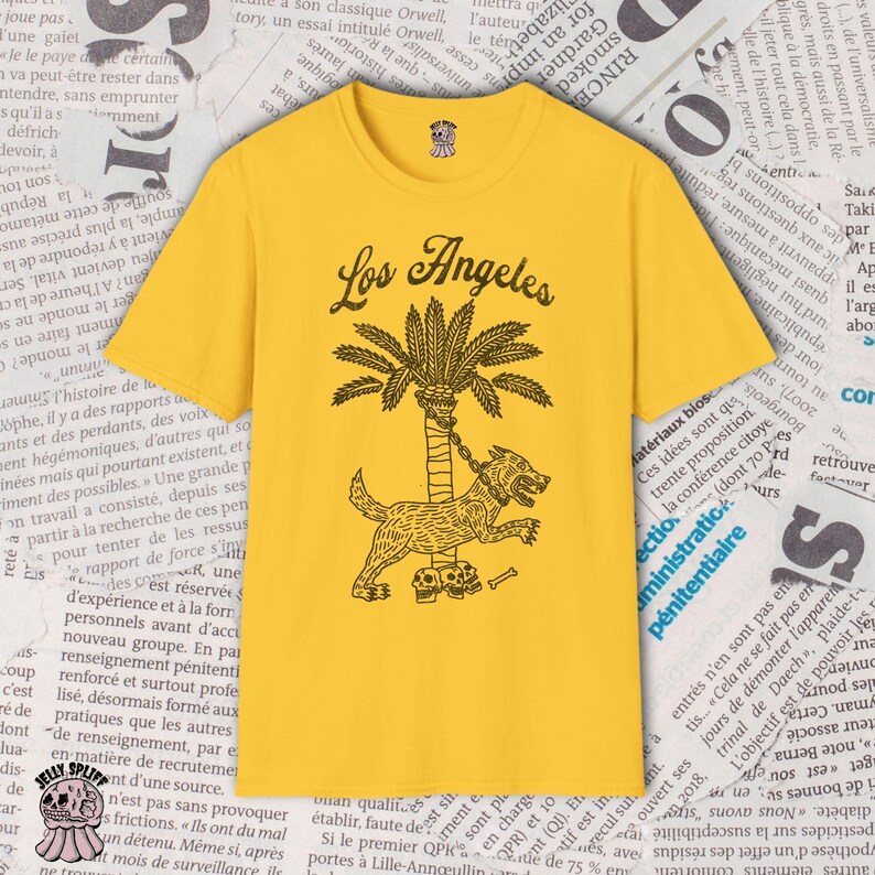May include: Mustard yellow t-shirt with the words "Los Angeles" above a palm tree and a dog illustration. The dog is chained to the tree and has skulls at its feet. The shirt has a round neck.