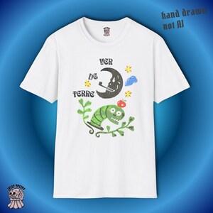 May include: White t-shirt with a whimsical graphic featuring a smoking skull moon, a green caterpillar, and the text "VER DE TERRE." The design includes stars, a cloud, and a red apple. The words "hand drawn not AI" are printed on the top right.
