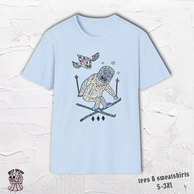 May include: Light blue t-shirt featuring a cartoon yeti skiing down a slope. The yeti has a surprised expression and is wearing skis. Above the yeti is a skull wearing a hat with wings. The shirt also has the text "tees & sweatshirts S-3XL".