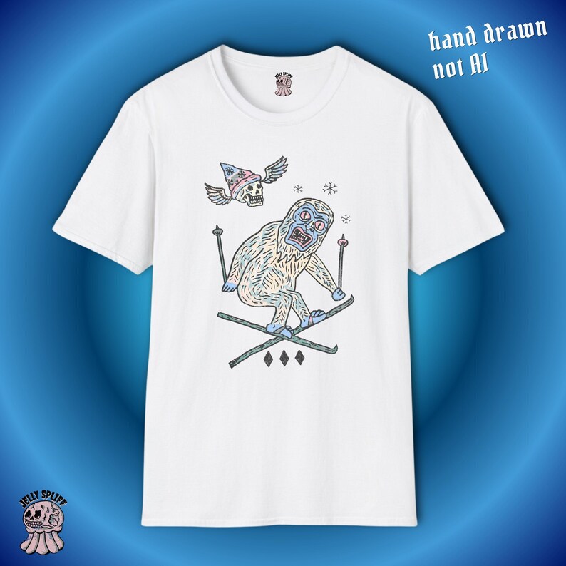 May include: White t-shirt featuring a cartoon yeti skiing, with a skull wearing a hat with wings above it. The text "hand drawn not AI" is printed on the top right of the shirt.