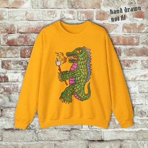 May include: Mustard yellow sweatshirt featuring a cartoon alligator roasting a marshmallow over a flame. The alligator is green with purple and blue accents. The text "hand drawn not AI" is in the upper right corner.