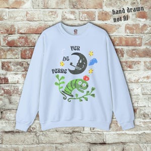 May include: A light blue sweatshirt with a whimsical design. The graphic includes a crescent moon with a skull face, a green worm, stars, and the words "VER DE TERRE". The text "hand drawn not AI" is also visible.