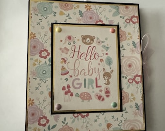 May include: A scrapbook with a floral pattern cover and a gold border. The front cover has a picture of a bear, a bunny, a turtle, and mushrooms. The text on the cover reads "Hello baby girl".