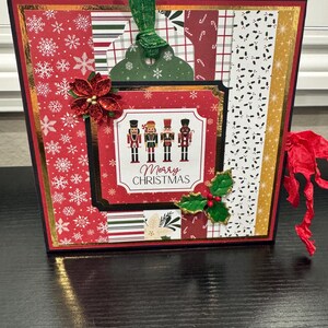 May include: A decorative Christmas gift bag with a red, gold, and black color scheme. The bag features a central panel with nutcracker illustrations and the words "Merry Christmas." Embellishments include a red poinsettia and green ribbon.