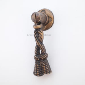 Op de afbeelding: Een bronzen kastgreep met een touwontwerp. De greep heeft een cirkelvormige achterplaat, een gedraaide touwachtige handgreep en een decoratieve kwast aan de onderkant. De hardware is ontworpen voor meubels en kasten.