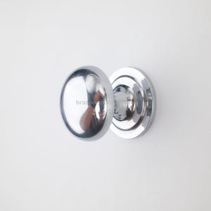 Puede incluir: Un pomo de puerta cromado brillante, con forma ovalada redondeada y base circular. El pomo refleja el entorno, creando un efecto de espejo. La ferretería está montada sobre una superficie blanca.