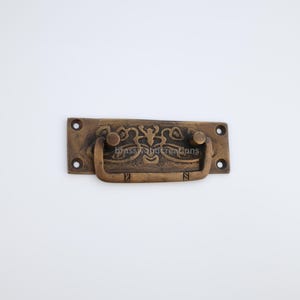 Peut inclure: Une poignée de tiroir en laiton avec un design orné et une forme rectangulaire. La poignée a deux petits boutons de chaque côté et une poignée encastrée au centre.