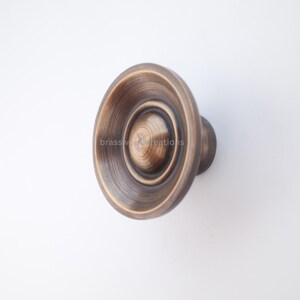 Puede incluir: Pomo de armario de bronce antiguo con dise&ntilde;o circular. El pomo presenta anillos conc&eacute;ntricos y un asa redondeada. El texto "brasswood creations" es visible en el pomo. Este herraje es adecuado para proyectos de mobiliario y decoraci&oacute;n del hogar.