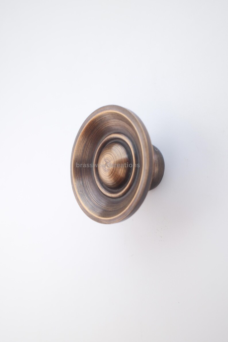 Puede incluir: Pomo de armario de bronce antiguo con dise&ntilde;o circular. El pomo presenta anillos conc&eacute;ntricos y un asa redondeada. El texto "brasswood creations" es visible en el pomo. Este herraje es adecuado para proyectos de mobiliario y decoraci&oacute;n del hogar.