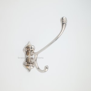 May include: A polished silver-toned metal wall hook featuring a dual-prong design with a long upper arm and a shorter lower arm. The base plate has four screw holes for mounting, and the text brasscreations is visible on the metal surface.