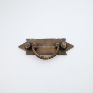May include: A brass drawer pull with a rectangular plate and a curved handle. The plate has a decorative design around the edges.