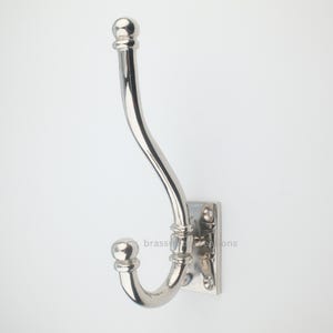 May include: A polished silver-toned metal wall hook featuring a double-pronged design with a long upper hook and a shorter lower hook. The base is a rectangular plate with four screw holes for mounting, and the text brassy creations is visible.