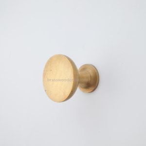 May include: A brass cabinet knob with a circular face and a short, cylindrical base. The knob is a warm gold color and is set against a white background. The text "brasswoodcreations" is visible on the knob.